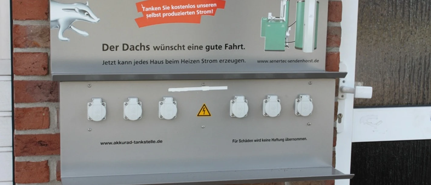 E-Bike-Ladesteckdosen, Andreas Fölling - Bad + Heizung Barrierefreie E-Tankstelle mit sechs weißen Steckdosen und einem Hinweisschild auf Backsteinwand.