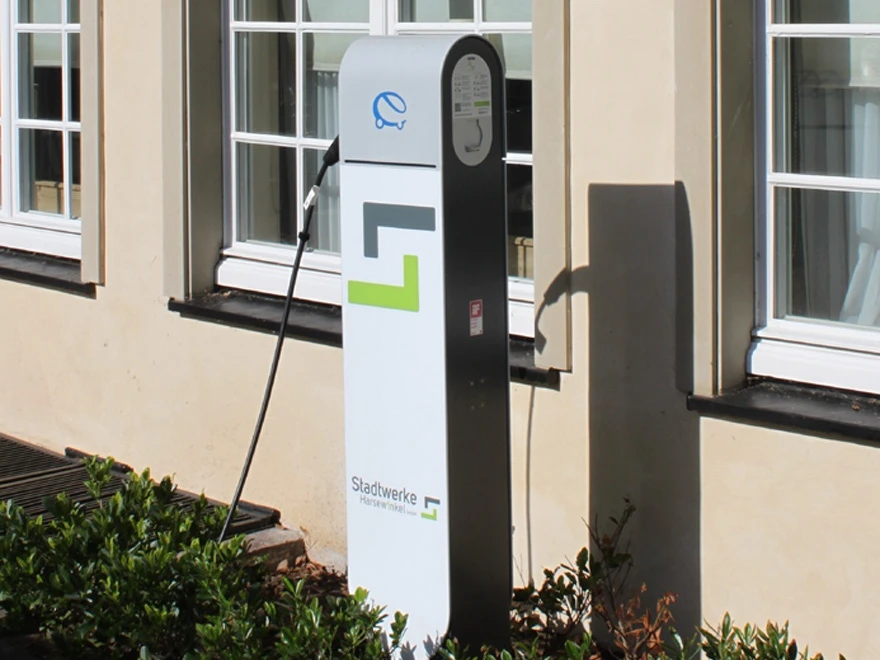 Kfz-Ladesäule an der Hauptstraße in Greffen Ladestation für Elektrofahrzeuge vor einem Gebäude mit weißen Sprossenfenstern und umgebenden Pflanzen.