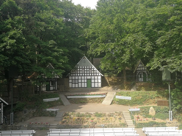 Freilichtbühne Kahle Wart, Bühne gesamt Eine Freilichtbühne mit Fachwerkhäusern im Hintergrund, umgeben von grünen Bäumen und Bänken.