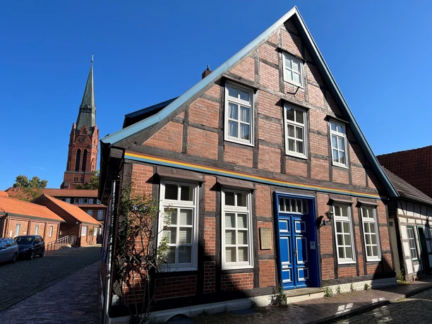 A half-timbered house with a blue door; in the background, a church tower rises against the clear blue sky.