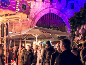 VeedelsAdvent on the Chlodwigplatz Festlich beleuchteter Chlodwigplatz bei Nacht. Die Severinstorburg im Hintergrund erstrahlt in violettem Licht, während zahlreiche Menschen im Vordergrund an einem kleinen Karussell stehen.Festively illuminated Chlodwigplatz at night. The Severinstorburg in the background shines in purple light, while numerous people stand at a small carousel in the foreground.