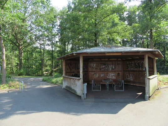 Holzhütte mit Graffiti und zwei Bänken auf asphaltiertem Weg im grünen Wald am Rundweg Steinhude.