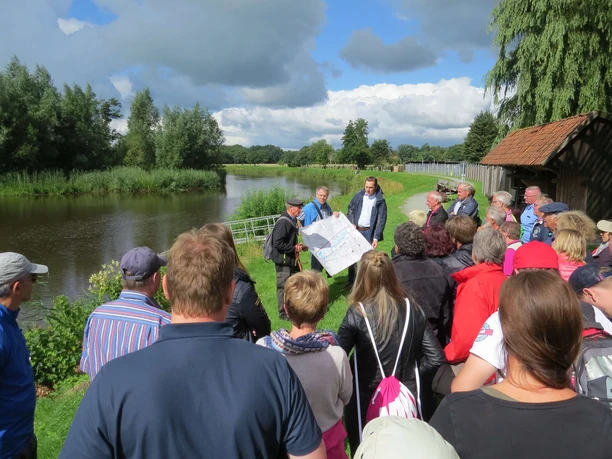 Deichwanderung am Aper Tief Eine Gruppe von Menschen steht auf einem grünen Deich nahe des Aper Tiefs unter bewölktem Himmel. Ein Mann in der Mitte hält eine Karte in die Höhe, während er den aufmerksam lauschenden Personen etwas erklärt.