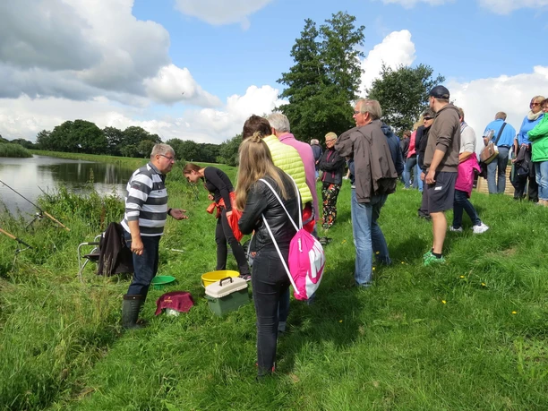 Naturerlebnis Aper Tief Verschiedene Menschen stehen am grünen Ufer eines friedlichen Gewässers und lauschen einer Vorführung.