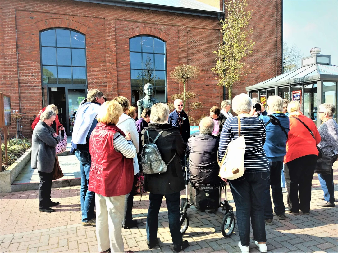 g-stef-hrung-zeitreise-augustfehn-apen Eine Gruppe von Menschen steht vor einem Backsteingebäude und hört einem Redner zu.
