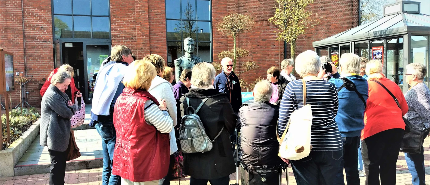 g-stef-hrung-zeitreise-augustfehn-apen Eine Gruppe von Menschen steht vor einem Backsteingebäude und hört einem Redner zu.