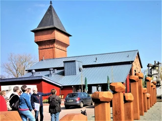 Eisenhütte-Puddelöfen-Augustfehn-Apen Eine Gruppe von Menschen steht vor einem historischen Backsteinbau mit einem großen Turm und modernen metallischen Akzenten. Ein blauer Himmel und zahlreiche Metallskulpturen sind im Vordergrund sichtbar.