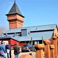 Eisenhütte-Puddelöfen-Augustfehn-Apen Eine Gruppe von Menschen steht vor einem historischen Backsteinbau mit einem großen Turm und modernen metallischen Akzenten. Ein blauer Himmel und zahlreiche Metallskulpturen sind im Vordergrund sichtbar.