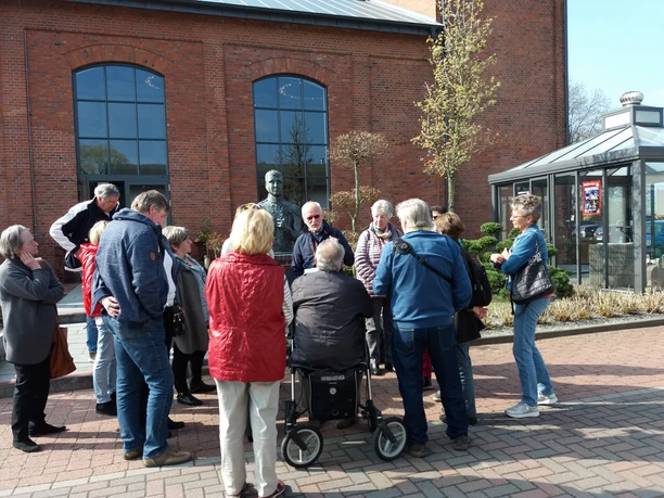 Gästeführung an der Eisenhütte Eine Gruppe von Personen nimmt an einer Führung vor einer historischen Backsteinfassade teil, darunter eine Metallbüste.
