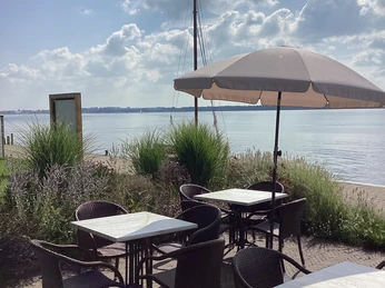 Terrasse an Anleger Insel Wilhelmstein Terrassenmöbel mit Blick auf den See und Segelmast, sanft beleuchtet von der Sonne, unter blauem Himmel.Terrace furniture with a view of the lake and sailing mast, gently illuminated by the sun, under a blue sky.Terrassemøbler med udsigt over søen og sejlmasten, blidt oplyst af solen under en blå himmel.Terrasmeubilair met uitzicht op het meer en de zeilmast, zachtjes verlicht door de zon, onder een blauwe hemel.