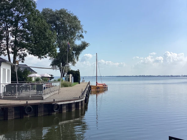 Jetty with sailing boats and trees in the background, on a calm stretch of water under a blue sky.