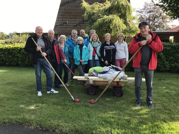 Aus dem Ammerländer Leben Eine Gruppe von elf älteren Menschen posiert lächelnd auf einer grünen Wiese mit einem Bollerwagen und Klootschießen-Stöcken.