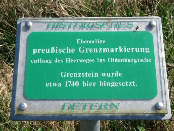 Grenzstein Holtgast Ein grünes Schild mit der Aufschrift "Ehemalige preußische Grenzmarkierung" steht in einer Wiese. Der Text erklärt, dass der Grenzstein entlang des Heerweges im Oldenburgischen etwa 1740 hier aufgestellt wurde. Das Schild ist mit "Historisches Deterner" gekennzeichnet.