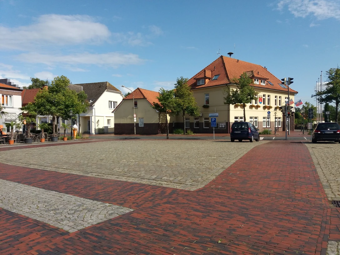rathausmarkt-apen Der Rathausmarkt Apen zeigt eine harmonische städtische Szenerie mit roten Pflastersteinen und Gebäuden im traditionellen Baustil. Ein blauer Himmel und grüne Bäume ergänzen das Bild.