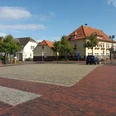 rathausmarkt-apen Der Rathausmarkt Apen zeigt eine harmonische städtische Szenerie mit roten Pflastersteinen und Gebäuden im traditionellen Baustil. Ein blauer Himmel und grüne Bäume ergänzen das Bild.