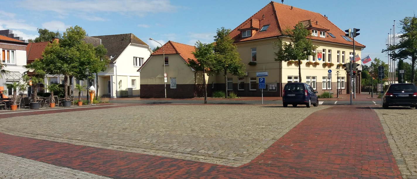 rathausmarkt-apen Der Rathausmarkt Apen zeigt eine harmonische städtische Szenerie mit roten Pflastersteinen und Gebäuden im traditionellen Baustil. Ein blauer Himmel und grüne Bäume ergänzen das Bild.