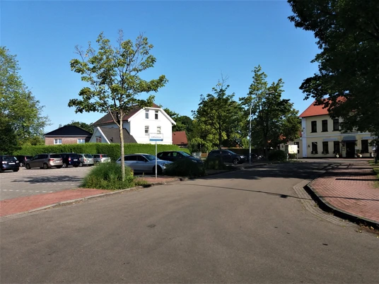 Viehmarktplatz-in-Apen Der Viehmarktplatz in Apen zeigt eine ruhige Straßenszene. Links parken Autos vor grünen Hecken und einem roten Dach. Im Hintergrund gibt es Bäume und der Himmel ist blau. Rechts steht ein hellgelbes Gebäude mit einem roten Dach.