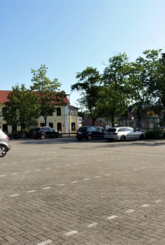 Aper Viehmarktplatz Ein Platz mit mehreren geparkten Autos. Im Hintergrund sind vereinzelte Bäume und ein gelbes Gebäude mit roten Dach zu sehen. Der Himmel ist klar und blau.