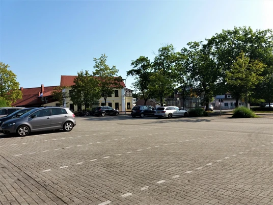 Aper Viehmarktplatz Ein Platz mit mehreren geparkten Autos. Im Hintergrund sind vereinzelte Bäume und ein gelbes Gebäude mit roten Dach zu sehen. Der Himmel ist klar und blau.