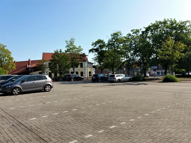 Aper Viehmarktplatz Ein Platz mit mehreren geparkten Autos. Im Hintergrund sind vereinzelte Bäume und ein gelbes Gebäude mit roten Dach zu sehen. Der Himmel ist klar und blau.