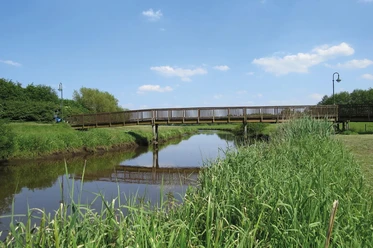 Große Süderbäke mit Heeren Wehren Brücke Eine Holzbrücke überspannt die ruhige Große Süderbäke, umgeben von grüner Natur unter blauem Himmel.