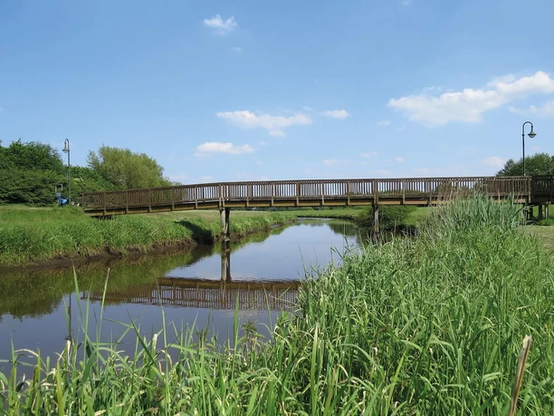 Große Süderbäke mit Heeren Wehren Brücke Eine Holzbrücke überspannt die ruhige Große Süderbäke, umgeben von grüner Natur unter blauem Himmel.