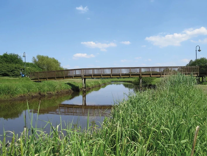 Große Süderbäke mit Heeren Wehren Brücke Eine Holzbrücke überspannt die ruhige Große Süderbäke, umgeben von grüner Natur unter blauem Himmel.