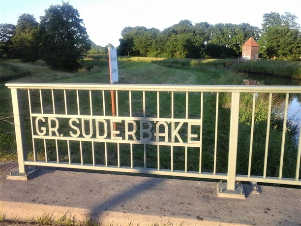 Brücke an der Süderbäke Metallbrücke über die Süderbäke mit Blick auf Grünflächen und ein kleines Backsteingebäude.