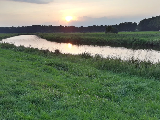 Godensholter Tief Das Bild zeigt einen sanften Flusslauf des Godensholter Tiefs, der malerisch durch grüne Wiesen fließt. Der Himmel ist von der untergehenden Sonne in warme Farben getaucht, die sich auf der Wasseroberfläche spiegeln. Eine friedliche, fast schon idyllische Landschaft breitet sich aus, die Ruhe und Naturverbundenheit ausstrahlt.