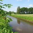 Nordlohkanal Ein ruhiger Kanal fließt durch eine grüne Landschaft, gesäumt von Bäumen und Wiesen unter blauem Himmel.