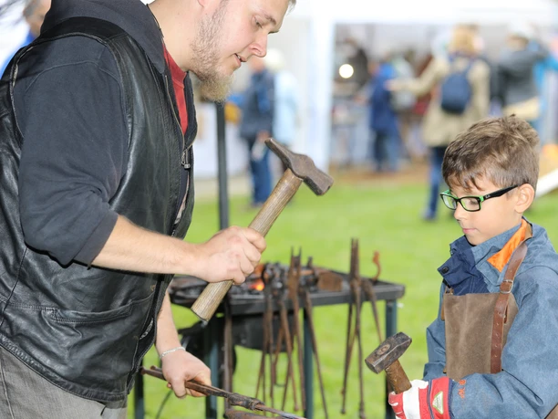 Herbstmarkt Ein Schmied zeigt einem Jungen, wie man mit Hammer und Amboss arbeitet, um Metall zu formen.
