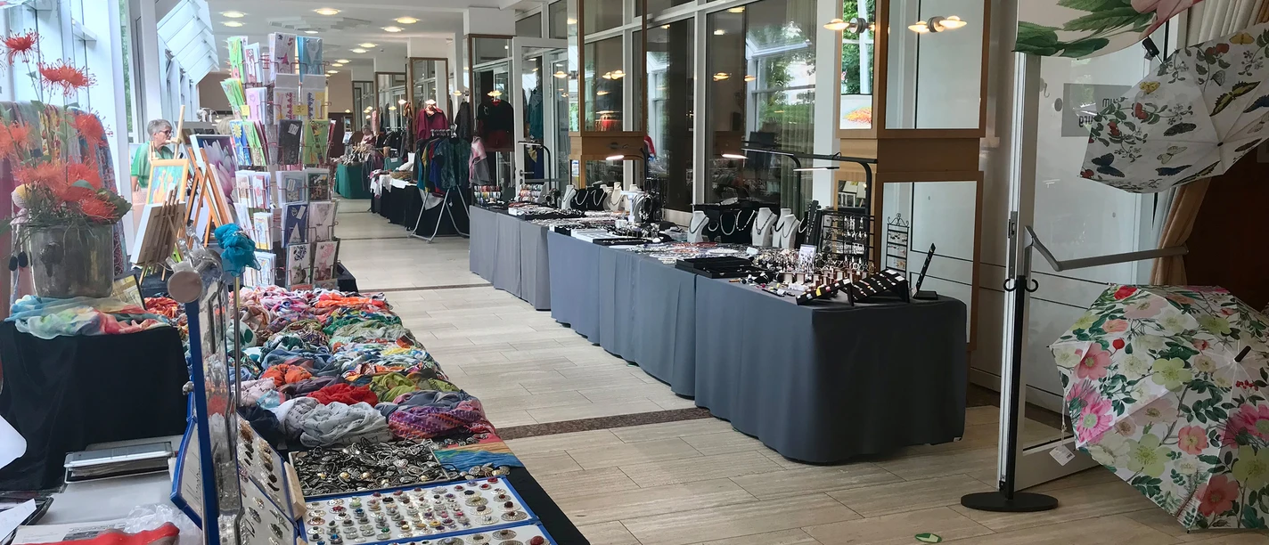 ausstellung.jpg An exhibition hall with a bright atmosphere features market stalls. Jewelry, colorful scarves, and decorative umbrellas are displayed on long tables. The floor-to-ceiling windows in the background let in plenty of natural light, giving the space a welcoming atmosphere.