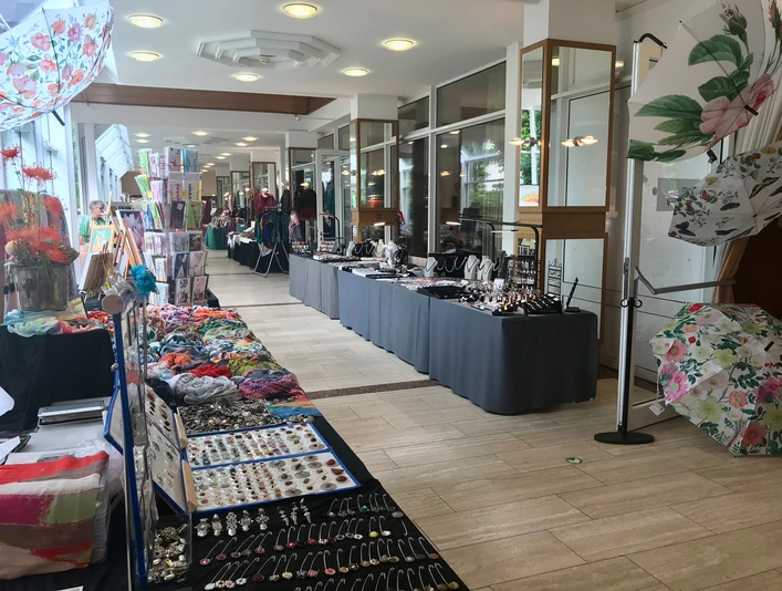 ausstellung.jpg Eine Ausstellungshalle mit einer hellen Atmosphäre zeigt Marktstände. Auf langen Tischen werden Schmuck, bunte Tücher und dekorative Schirme präsentiert. Die Fensterfront im Hintergrund sorgt für viel Tageslicht, das den Räumlichkeiten eine einladende Atmosphäre verleiht.