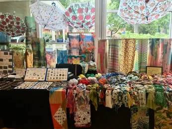 ausstellung-schals.jpg Eine bunte Kollektion von Schals ist auf einem Marktstand drapiert, umgeben von farbenfrohen Textilmustern und dekorativen Accessoires.