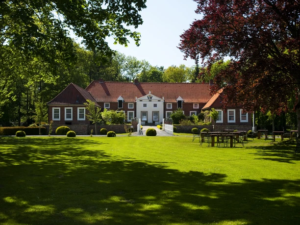Een bakstenen landgoed met een rood dak, omgeven door een groen park en oude bomen in de zon.