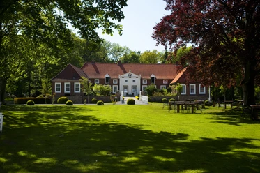 Familotel Gut Landegge Backstein-Gutshof mit rotem Dach, umgeben von grüner Parkanlage und alten Bäumen im Sonnenschein.