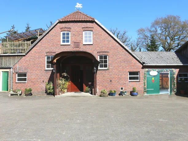 Der Ammerländer Kerzenstall von außen Rustikales rotes Backsteingebäude mit gepflastertem Hof, Holzeingang und einem grünen Tor.