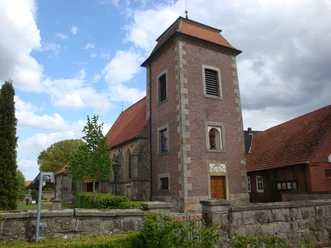 St.-Johannes-Kirche in Elbergen Backsteinkirche mit quadratischem Turm, roten Ziegeldächern und steinerner Mauer im Vordergrund.