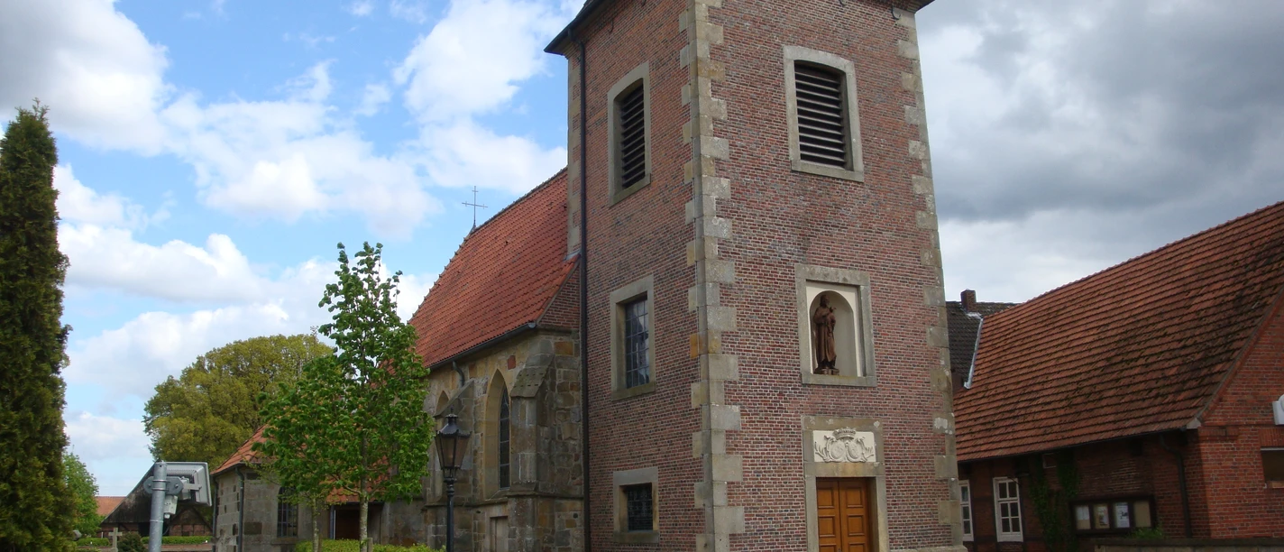 St.-Johannes-Kirche in Elbergen Backsteinkirche mit quadratischem Turm, roten Ziegeldächern und steinerner Mauer im Vordergrund.