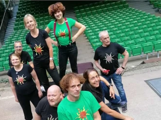 spekspek-web.jpg Eine fröhliche Gruppe steht auf einer Treppe vor einer Reihe von grünen Sitzplätzen. Alle tragen T-Shirts mit einem auffälligen Spinnenmotiv.