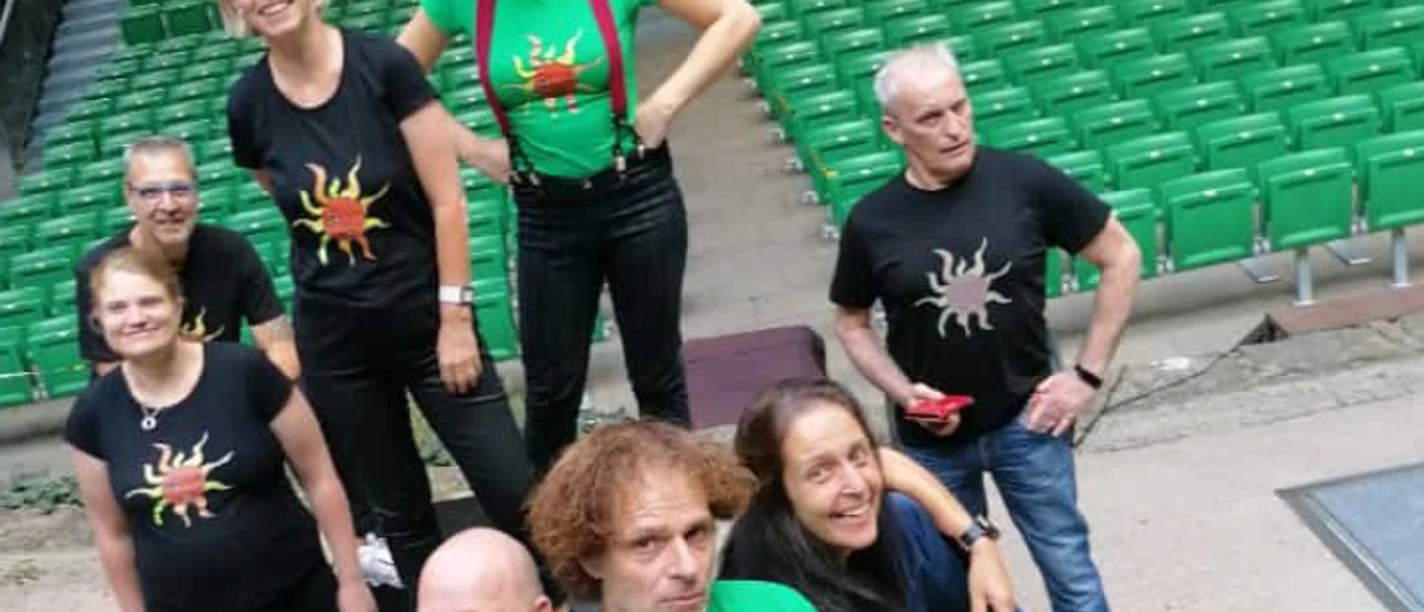 spekspek-web.jpg Eine fröhliche Gruppe steht auf einer Treppe vor einer Reihe von grünen Sitzplätzen. Alle tragen T-Shirts mit einem auffälligen Spinnenmotiv.