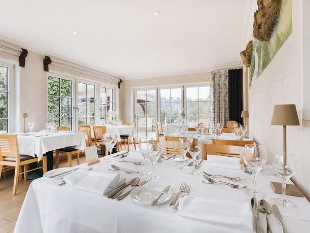 Bright restaurant with large windows, tables laid with white tablecloths, glasses and cutlery, wooden furniture and a quiet atmosphere.