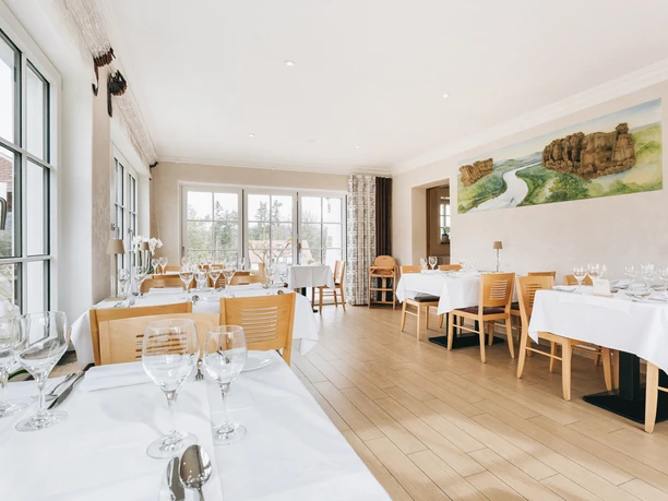 Bright restaurant with wooden floor, covered tables and large windows; a landscape painting on the wall.