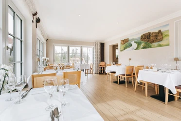 Restaurant im Laasenhof Bright restaurant with wooden floor, covered tables and large windows; a landscape painting on the wall.