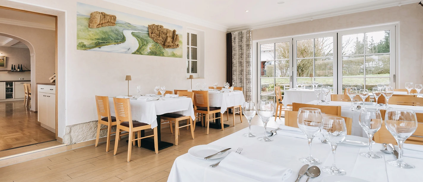 Licht restaurant met houten vloer, gedekte tafels en grote ramen met uitzicht op het groen; aan de muur hangt een landschapschilderij.