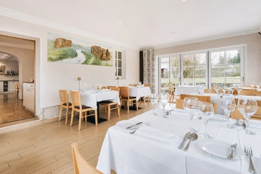 Restaurant im Laasenhof Helles Restaurant mit Holzboden, gedeckten Tischen und großen Fenstern mit Blick ins Grüne; an der Wand hängt ein Landschaftsbild.