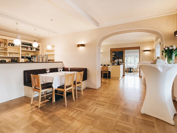 Bright restaurant with wooden floor, set table and chairs, bar area in the background, white tablecloth, tulip vase on bar table.