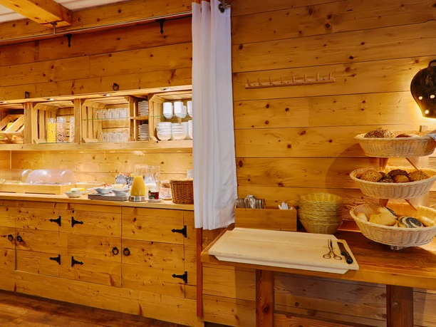 Gasthof Sennerhütte Frühstücksbuffet in einem rustikalen Raum mit Holzwänden, gefüllt mit Brotkörben, Saft, Geschirr und Besteck, beleuchtet von einer warmen Lampe.