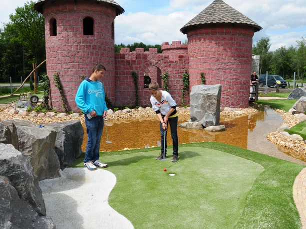 Adventure Golfanlage Dankern Zwei Personen spielen Minigolf auf einer naturnah gestalteten Anlage vor einer roten Burgkulisse.