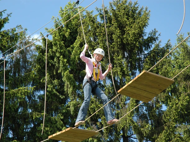Person mit Klettergurt und Helm balanciert im Hochseilgarten zwischen Holzplatten über Seilen.
